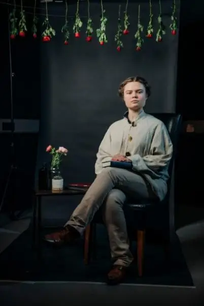 A female cast member sat on a chair with her legs crossed. There are roses hung above her.