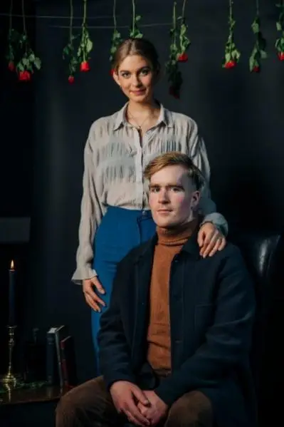 A male sat on a chair and a female stood behind him with a hand on his shoulder